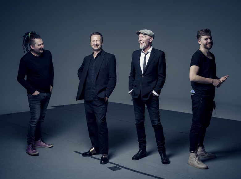 Czterech roześmianych mężczyzn stoi w rzędzie na tle szarej ściany. Ubrani są na ciemno, mają ręce w kieszeniach. To członkowie zespołu Voo Voo.