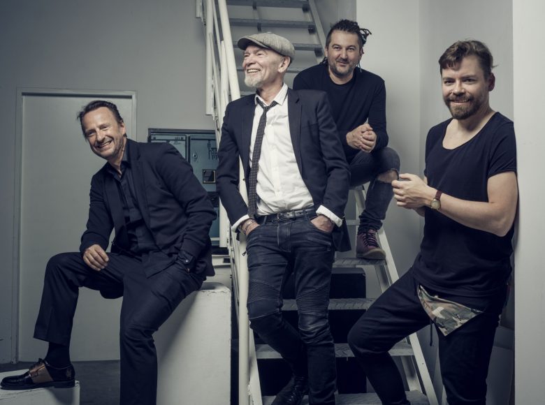 Czterech mężczyzn na zapleczu sceny. Część z nich siedzi na metalowych schodkach prowadzących do góry, reszta stoi wokół. Są w dobrym nastroju, uśmiechają się. To członkowie zespołu Voo Voo.