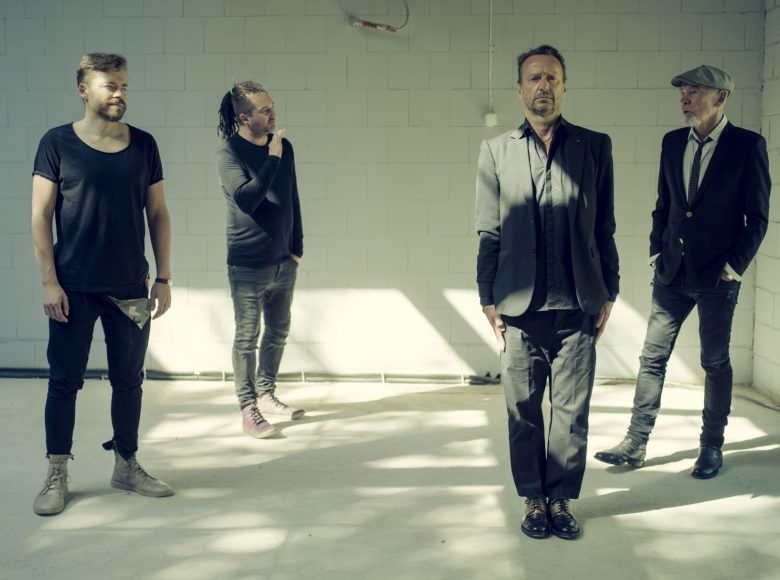 Czterech mężczyzn ubranych na czarno stoi w rozsypce w surowym murowanym wnętrzu budynku. Oświetla ich światło wpadające przez okno. Sprawiają wrażenie zamyślonych. To członkowie zespołu Voo Voo.