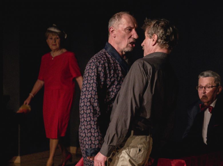 Scena ze spektaklu "Chłopcy". Dwóch aktorów stoi na przeciw siebie, kłócą się. Na nimi siedzi mężczyzna w marynarce i muszce. Obok przechodzi kobieta w czerwonej sukience.