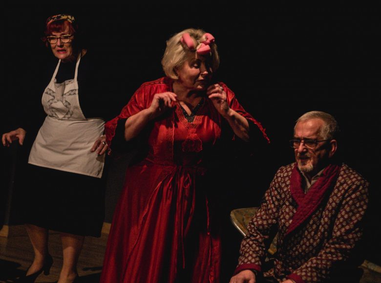 Scena ze spektaklu "Chłopcy" Kobieta w czerwonym szlafroku i papilotach na głowie rozmawia z mężczyzną, który siedzi na ławce. obok nich skrada się pokojówka w czarnej sukience i białym fartuszku.