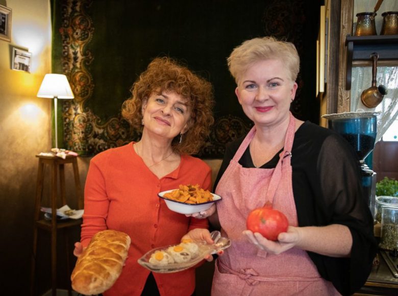 Dwie uśmiechnięte kobiety trzymają w rękach różne dania. W tle wnętrze restauracji.