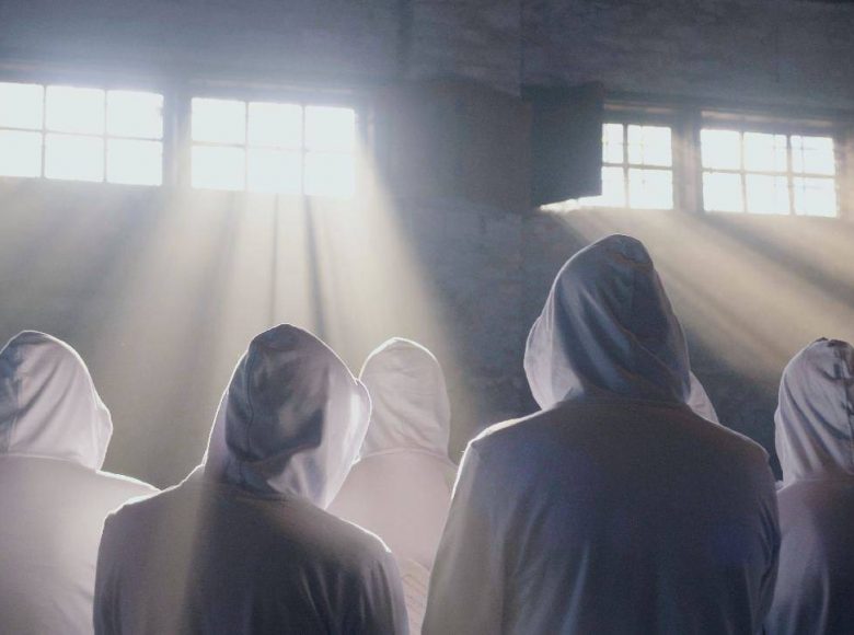 Wnętrze budynku. Kilka stojących tyłem postaci. Ubrane są w białe bluzy, mają na głowach kaptury. Ich sylwetki oświetla jasne promienie słońca wpadające przez okno.