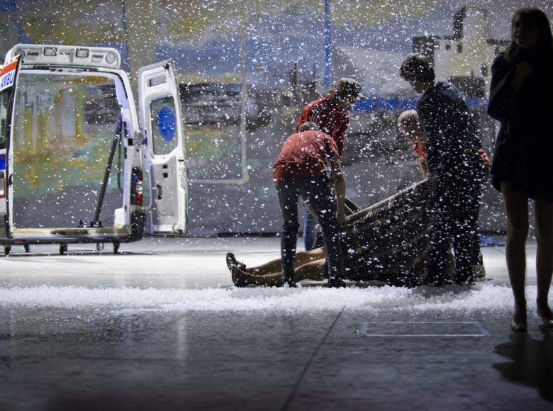Sylwetki grupy postaci po prawej stronie. 4 osoby podnoszą leżącą postać, widać tylko nogi. Po prawej karetka z otwartymi tylnymi drzwiami. Nad całością sceny unoszą się płatki śniegu.