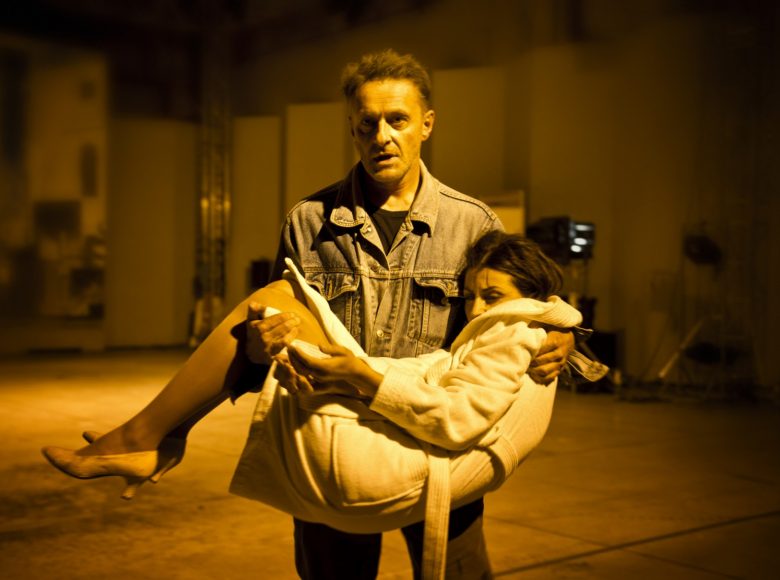 Mężczyzna trzyma w ramionach kobietę ubraną w biały płaszcz za kolano. Scena oświetlona na żółto.