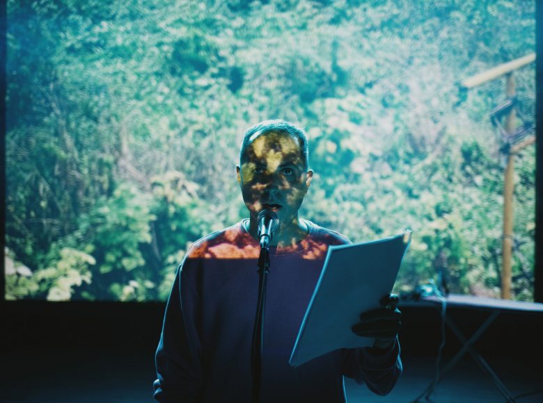 Mężczyzna stoi przy mikrofonie. W dłoni trzyma plik kartek, patrzy przed siebie. Twarz częściowo przysłonięta obrazem roślinności, który jest wyświetlany za postacią.