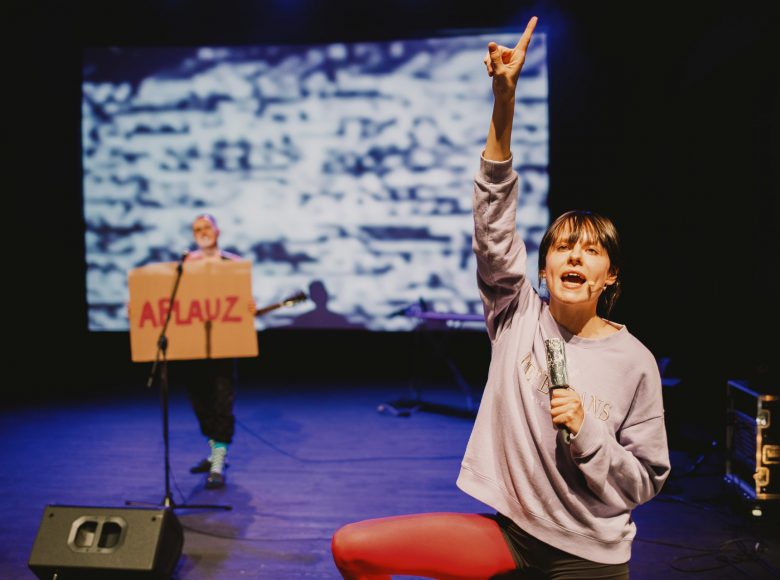 Dwie kobiety stojące na scenie. Kobieta po prawej stoi bliżej, jedną rękę ma uniesioną nad siebie, w drugiej trzyma rolkę srebrnej folii. Kobieta w tle trzyma transparent z napisem „Aplauz”.