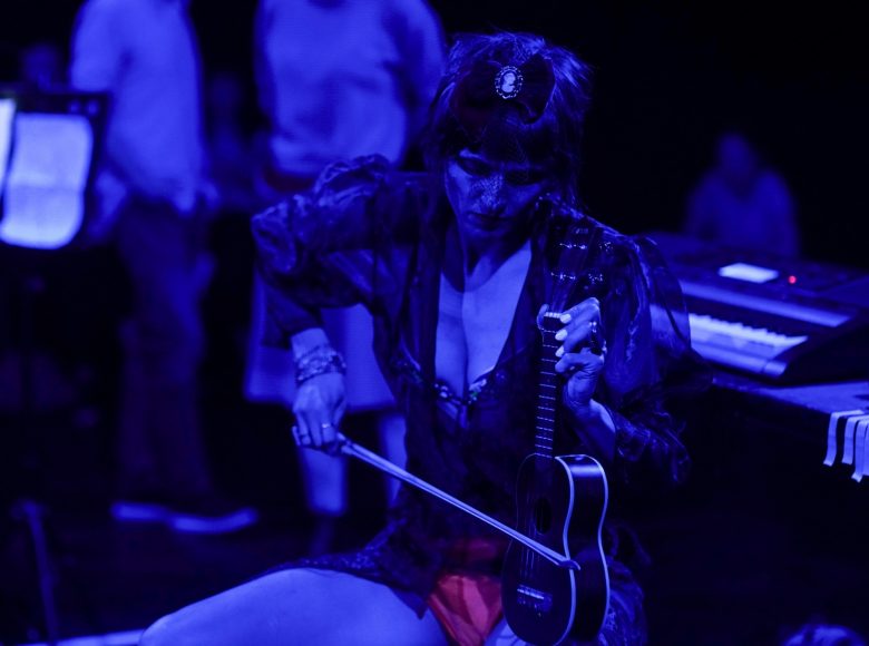 Kobieta w skąpym, półprzezroczystym stroju, oświetlona ciemnoniebieskim światłem. Siedzi, trzyma małą gitarę opartą na kolanie, gra na niej za pomocą smyczka.