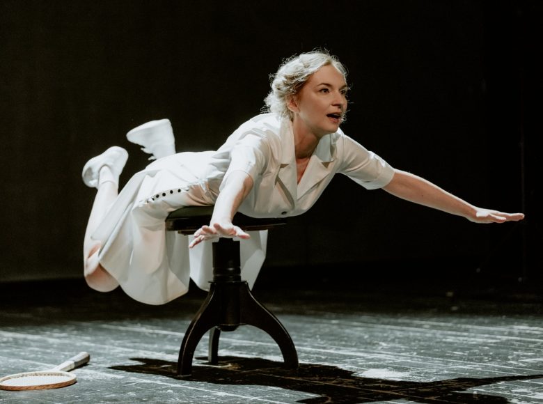 Kobieta w białym stroju do gry w tenisa z epoki międzywojennej. Leży na niskim czarnym stołku, ręce i nogi unosząc w powietrzu.