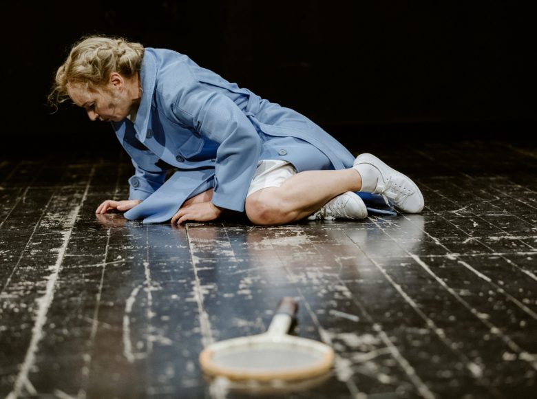 Kobieta w jasnoniebieskim płaszczu. Leży na ziemi, skulona, podparta na jednej ręce. Przed nią rakieta tenisowa.