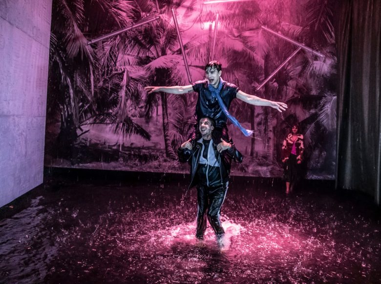 Dwóch mężczyzn w dynamicznej scenie. Jeden siedzi drugiemu na ramionach, mają rozłożone ręce, śmieją się. Dookoła nich pada deszcz, w tle obrazy liści palmowych oświetlone na różowo.