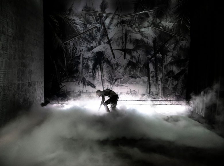 Czarno-białe zdjęcie. Częściowo niewidoczna sylwetka osoby, pochylonej do ziemi. Nad ziemią unoszą się kłęby gęstego białego dymu.