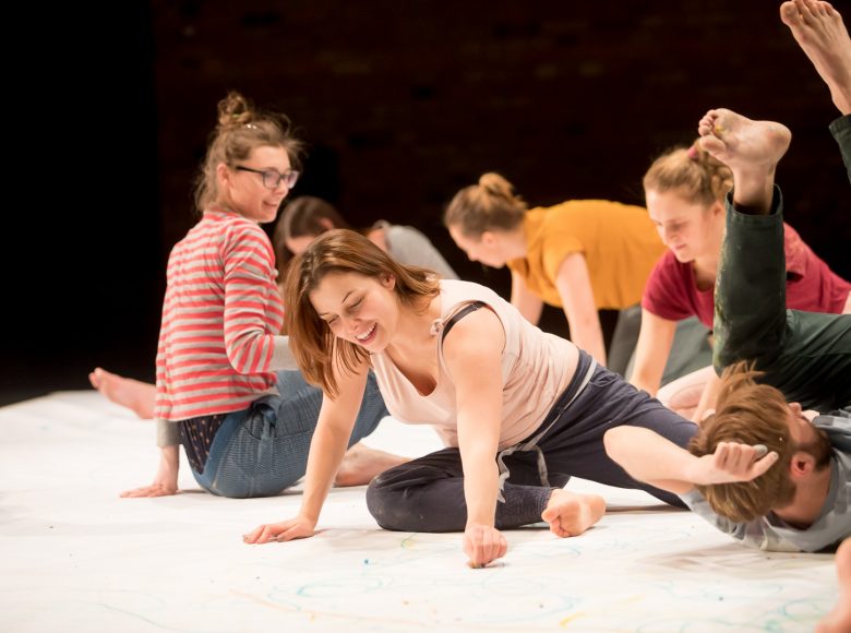 Grupa dzieci i dorosłych klęczy na ziemi. Kolorowymi pastelami wykonują rysunki na białym arkuszu rozłożonym na podłodze.