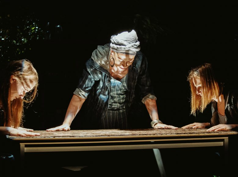 Trzy kobiety pochylone nad stołem. Dłonie mają położone przed sobą, na stole, twarze oświetlone od dołu.