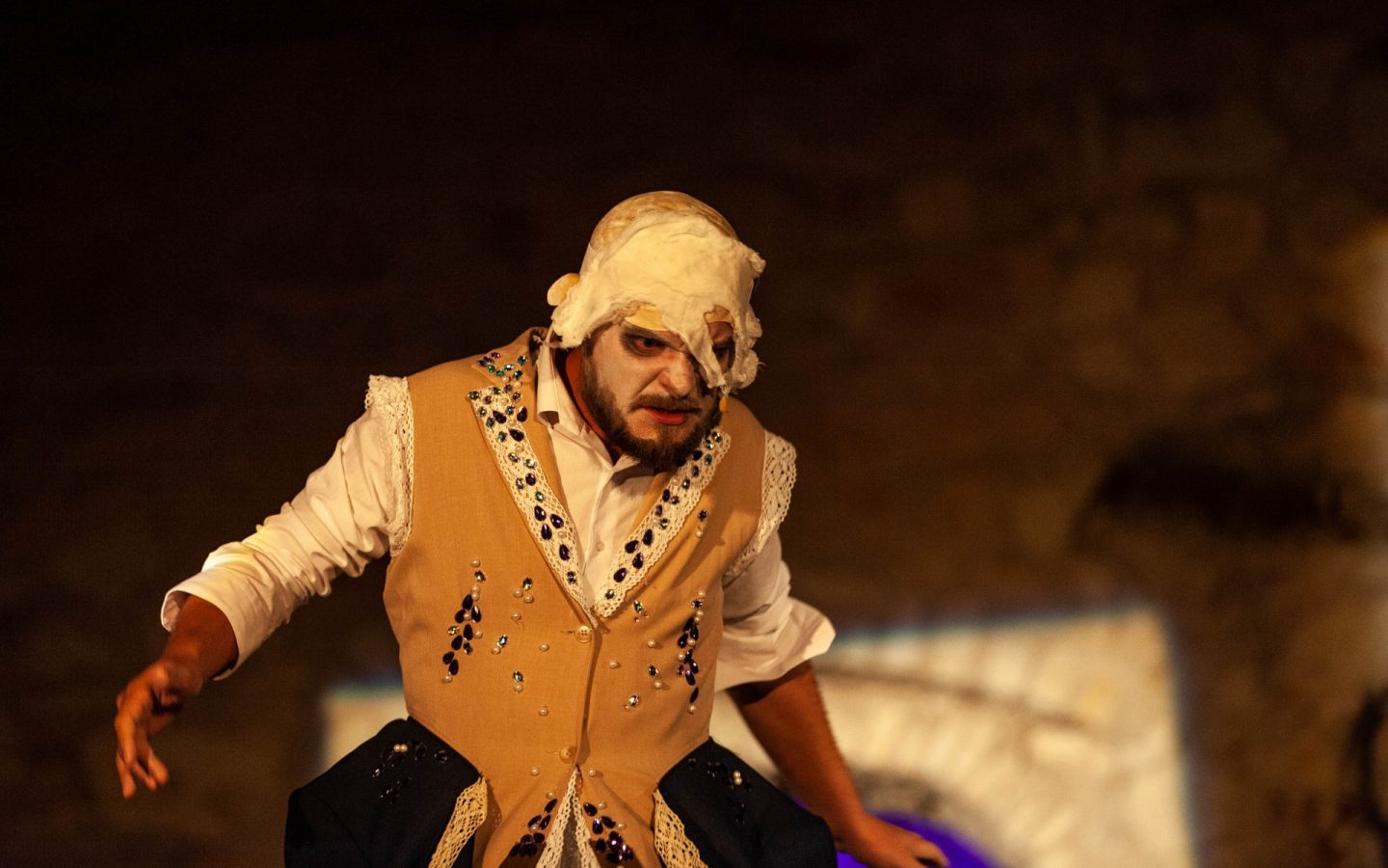 Mężczyzna na zdjęciu, jest ubrany w białą koszulę oraz kamizelkę garniturową z doszytą u dołu obszerną spódnicą. Jego twarz i głowę przysłania biała maska. Twarz jest pomalowana na biało a oczy podkreślone ciemnym kolorem. Kostium jest ubogacony wieloma błyszczącymi koralikami. Mężczyzna jest w ruchu, zmierza przed siebie poruszając rękami. Jego mimika wyraża grymas zacietrzewienia.