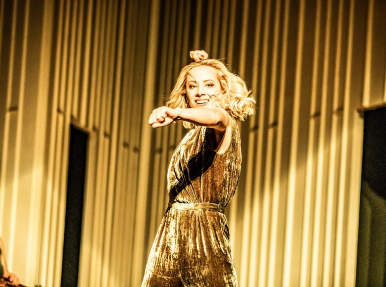 Kobieta w dynamicznej pozie. Ubrana w złotą sukienkę, jedną rękę ma uniesioną przed sobą, włosy rozwiane, uśmiecha się. Tło złote.