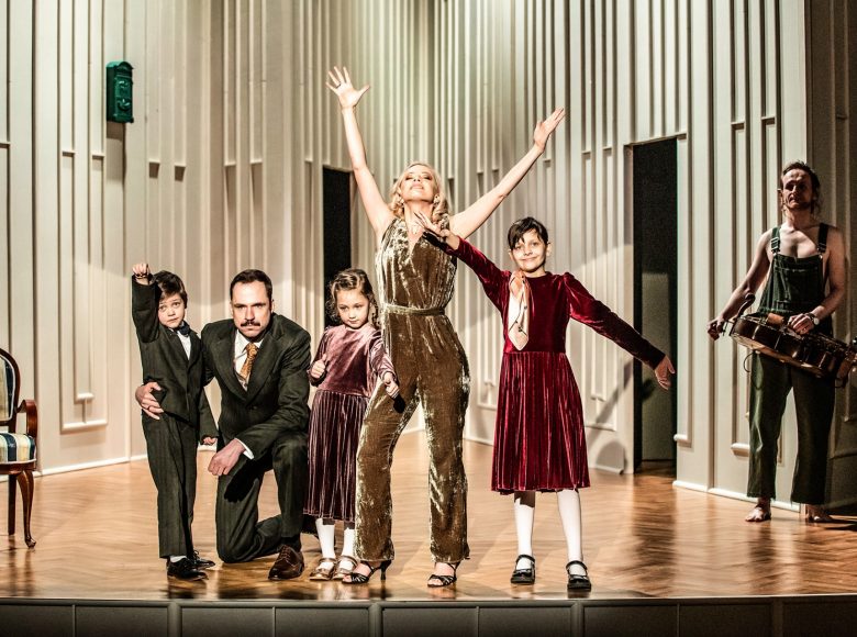 Scena grupowa. Kobieta w złotym stroju stoi centralnie, z uniesionymi nad siebie rękami. Dookoła niej trójka dzieci, dwie dziewczynki mają też uniesione ręce. Obok chłopca klęczy mężczyzna, obejmując go w pasie.