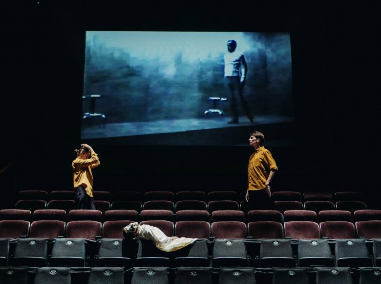 Scena ze spektaklu " Margules. Nieobecność". Aktorzy w żółtych koszulach znajdują się na miejscach przeznaczonych dla widowni. Za nimi na ekranie wyświetla się film.