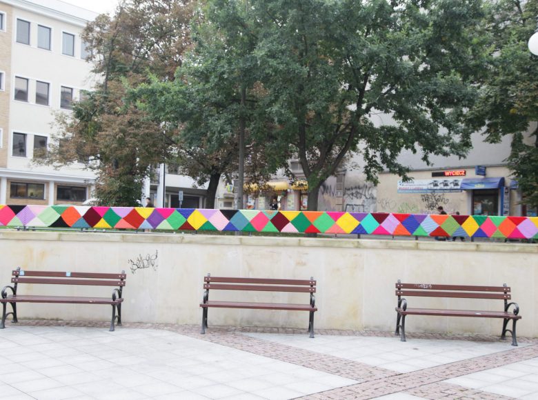 Przestrzeń przed Centrum Kultury w Lublinie. Murek owinięty jest kolorową ozdoba wykonana z włóczki. Pod murkiem stoja trzy ławki. W tle znajdują się sklepy i budynki biurowe.