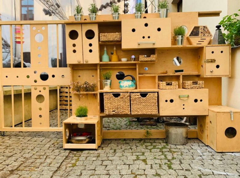 Zdjęcie przedstawia stojący na kostce brukowej kredens z naturalnej sklejki. Ma rozmaite półki i szafki z nawierconymi otworami w różnych rozmiarach. Na niektórych półkach stoją koszyki, metalowe doniczki z ziołami, domek dla owadów.