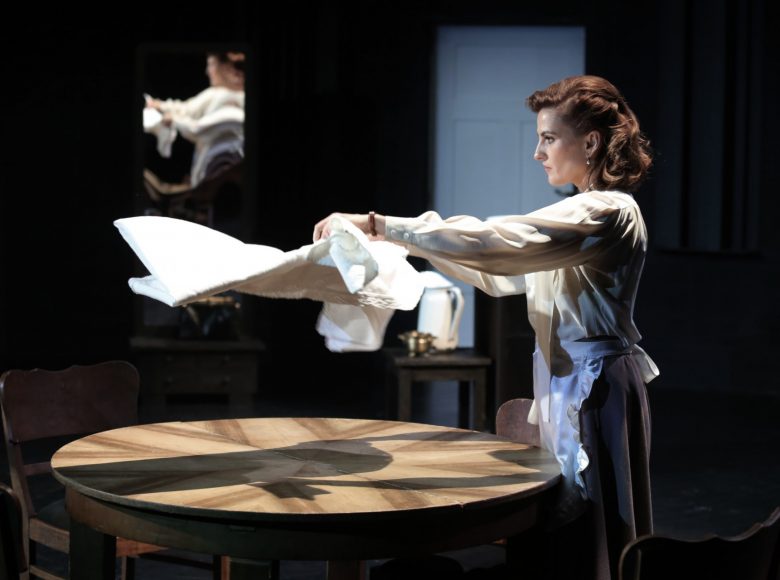 Elegancka kobieta, ubrana w białą bluzkę, fartuszek zawiązany w pasie. Rozkłada na drewnianym, okrągłym stole biały obrus.