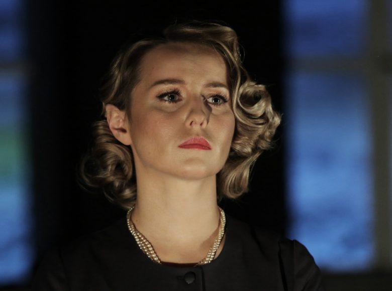 Zbliżenie na twarz eleganckiej kobiety. Blond włosy ułożone w loki. Na szyi ma trzy sznury pereł. Wydaje się smutna.