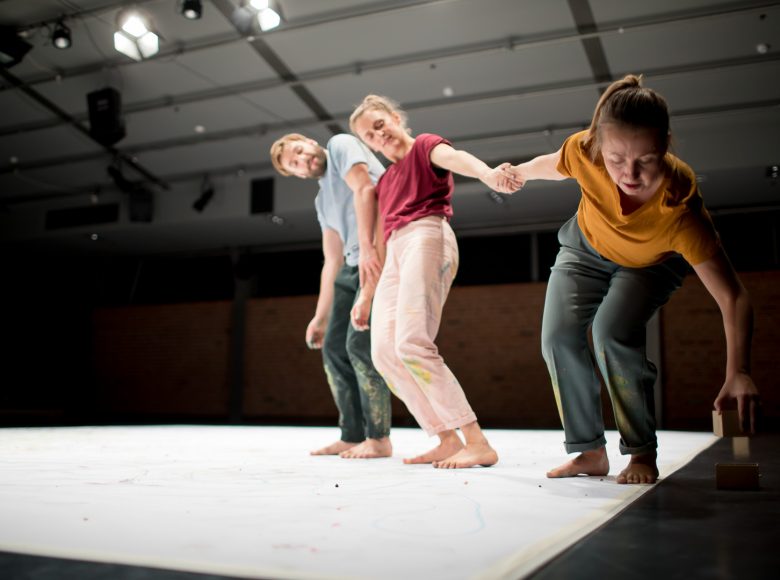 Grupa trzech osób na scenie. Trzymają się za ręce. Jedna z osób pochyla się żeby podnieść drewniany klocek.