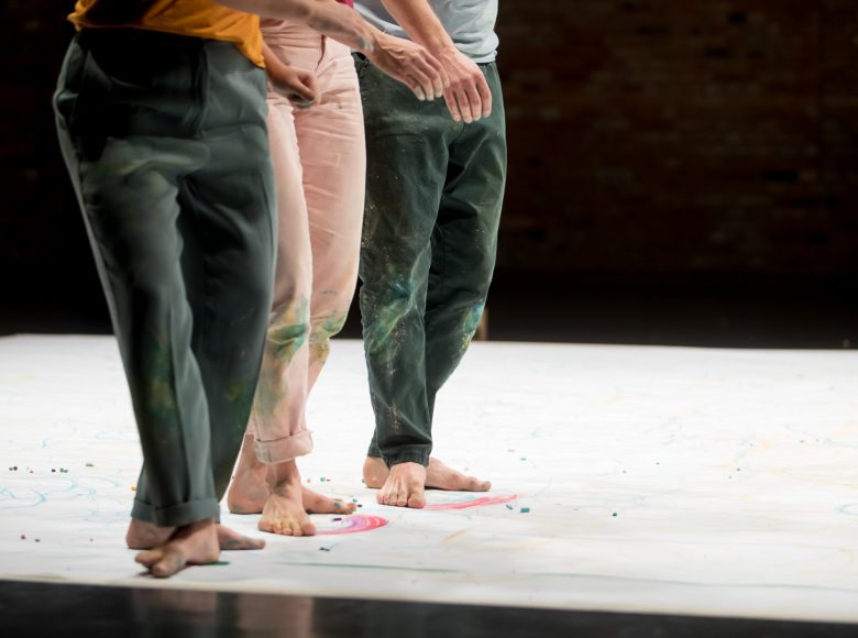 Trzy osoby stoją na białym arkuszu, rozłożonym na podłodze. Bosymi stopami rozmazują okruszki z pasteli.