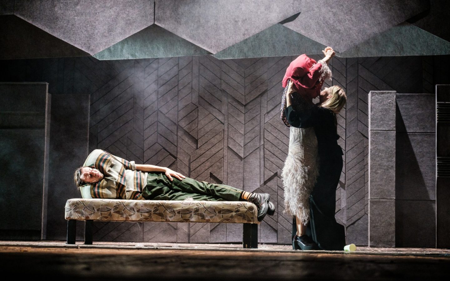 Grupa osób w przestrzeni pokoju, ściany i sufit ciemno szare, miękkie. Po lewej mężczyzna leży na niskim łóżku. Po prawej kobieta w wieczorowej sukni, druga kobieta zdejmuj jej przez głowę czerwony element ubioru.