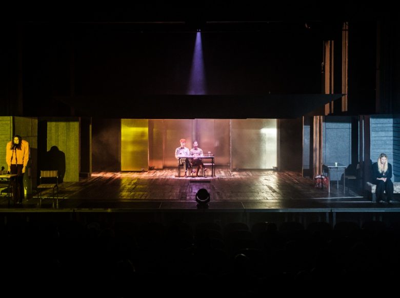 Szeroki kadr na scenę teatralną podzieloną na trzy sekcje. Każda sekcja oświetlona innym kolorem. Po lewej, w zielonym świetle stoi kobieta. Na środku, w żółtym świetle, dwóch mężczyzn siedzi za stołem. Po prawej, w niebieskim świetle, mężczyzna i kobieta siedzą na niskim łóżku.