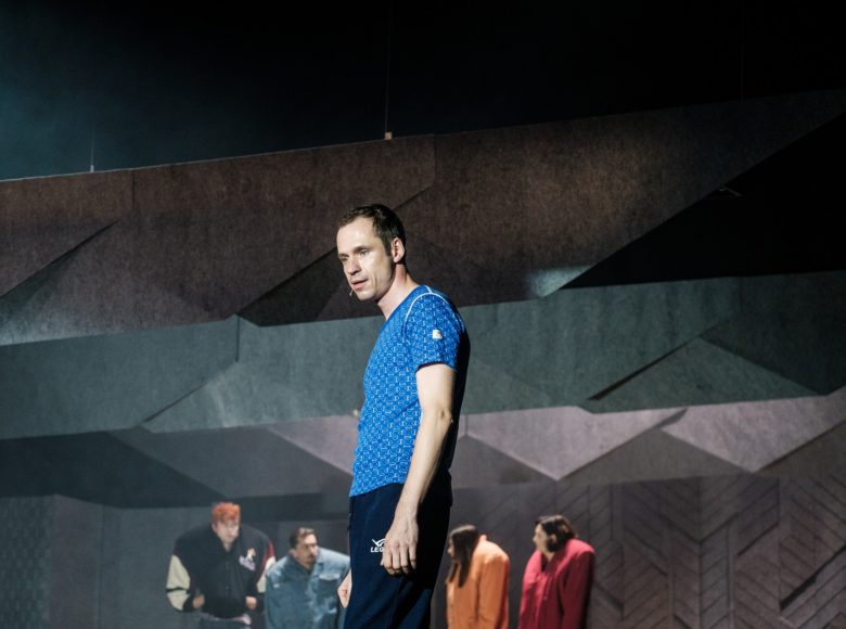 Mężczyzna na pierwszym planie, patrzy przed siebie. W tle grupa postaci z wysokimi, sztucznie uniesionymi ramionami.