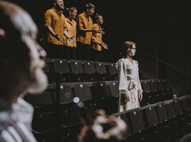 Scena ze spektaklu Margules. Nieobecność. Na pierwszym planie znajduje się kobieta w białej sukience. Ma rozpuszczone włosy i pomalowana na biało twarz. Za nią stoi grupa ludzi w żółtych koszulach.