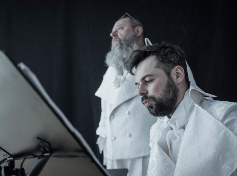 Dwóch mężczyzn w białych strojach. Na pierwszym planie młodszy, ma przed sobą pulpit, czyta. Starszy stoi w tle, patrzy przed siebie.