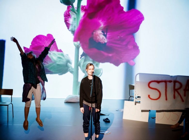 Dwie kobiety w szerokim kadrze. Kobieta po prawej w wyskoku, z rękami uniesionymi nad siebie. Druga stoi w środku zdjęcia, przed nią mikrofon. W tle wyświetlone różowe kwiaty malw. Po prawej łóżko szpitalne i karton z napisem „Strajk”.
