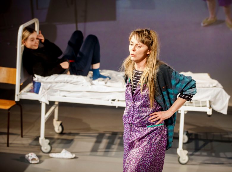 Na pierwszym planie kobieta w fartuchu w drobne kwiaty, ręce ma oparte na biodrach. W tle, częściowo niewyraźne, stoi łóżko szpitalne, na którym leży druga kobieta.