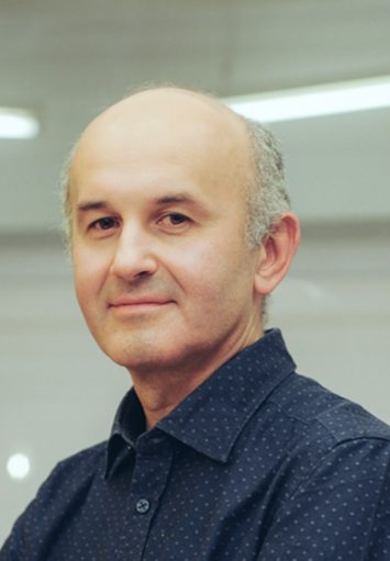 W industrialnej oświetlonej jarzeniówkami przestrzeni stoi łysy mężczyzna w średnim wieku. Ma na sobie ciemną koszulę i lekko się uśmiecha. Ręce ma założone na piersi.