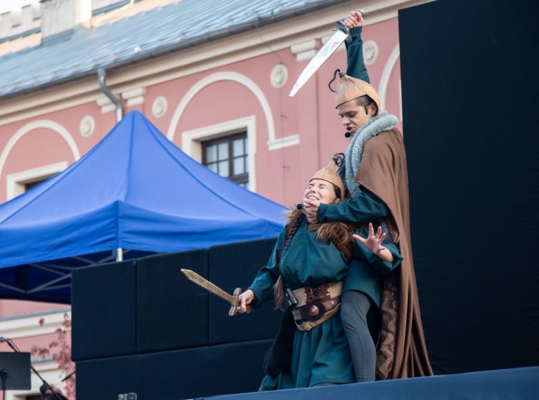 Aktorzy na scenie. Mężczyzna ubrany w ciemny kostium, hełm i brązową pelerynę atakuje mieczem klęcząca kobietę. Na twarzach aktorów wyrażają gniew i strach.