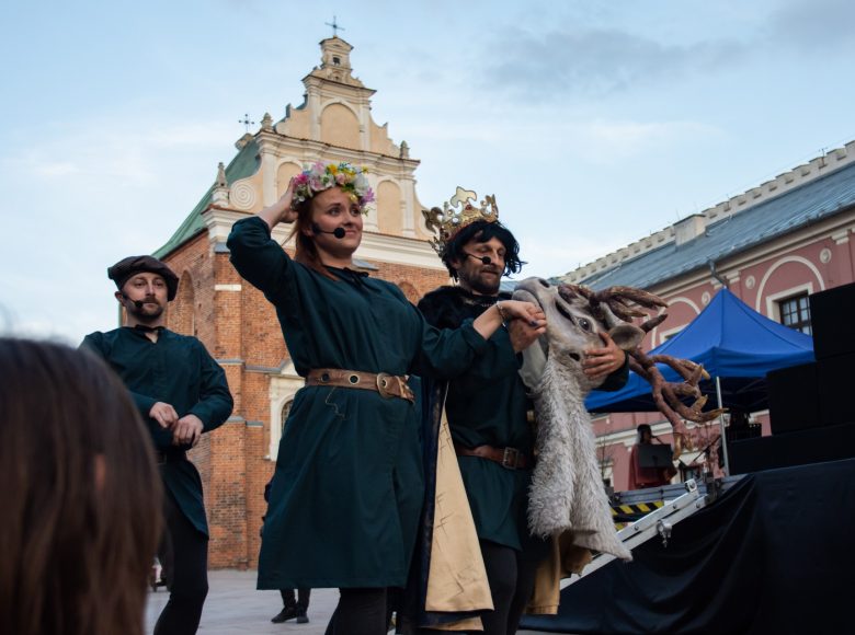 Aktorzy w trakcie przedstawienia. Mężczyzna i kobieta idą, trzymając się za ręce. Kobieta nosi na głowie wianek a mężczyzna koronę.