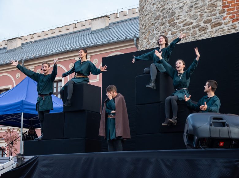 Aktorzy w identycznych kostiumach są ustawieni na scenie w różnych ekspresyjnych pozach. Ich miny wyrażają radość. Na środku sceny stoi mężczyzna w pelerynie. Zasłania twarz ręką, jest zmartwiony.