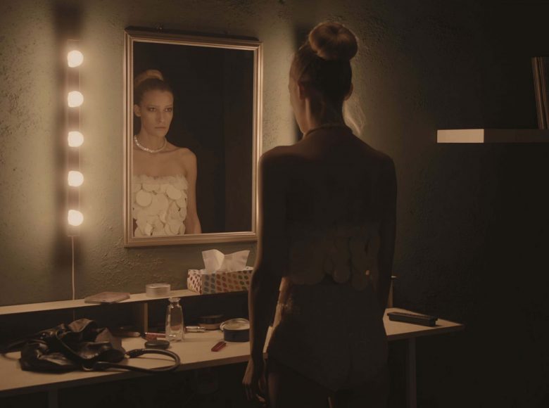 [ Kadr z filmu. Modelka w skąpym stroju stoi przed lustrem w garderobie. Bacznie przygląda się swojemu odbiciu. ]