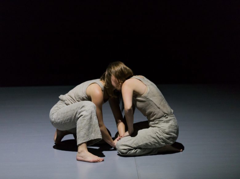 Dwie bose tancerki, w identycznych kostiumach klęczą na ziemi i wpatrują się sobie w oczy. Tło jednolite ciemne.