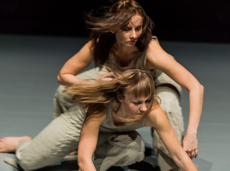 Dwie tancerki w identycznych, sasnych, luźnych kostiumach wykonują choreografię. Mają rozpuszczone, rozwiane włosy.