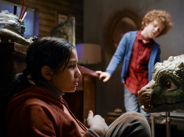 Kadr z filmu. Dziewczynka siedzi na podłodze w pokoju. Na przeciwko niej stoi mały, zielony smok i się w nią wpatruje. Z tyłu stoi chłopiec w czerwone koszuli i niebieskiej bluzie.