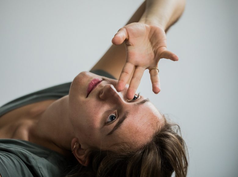 Zdjęcie. Tancerka odchyla swoje ciało do tyłu, rękę przykłada do twarzy. Tło jasne, jednolite.