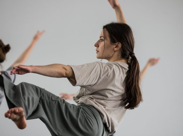 Grupa tancerek w luźnych, szarych kostiumach wykonuje skomplikowane figury choreograficzne. Tło jasne, jednolite.