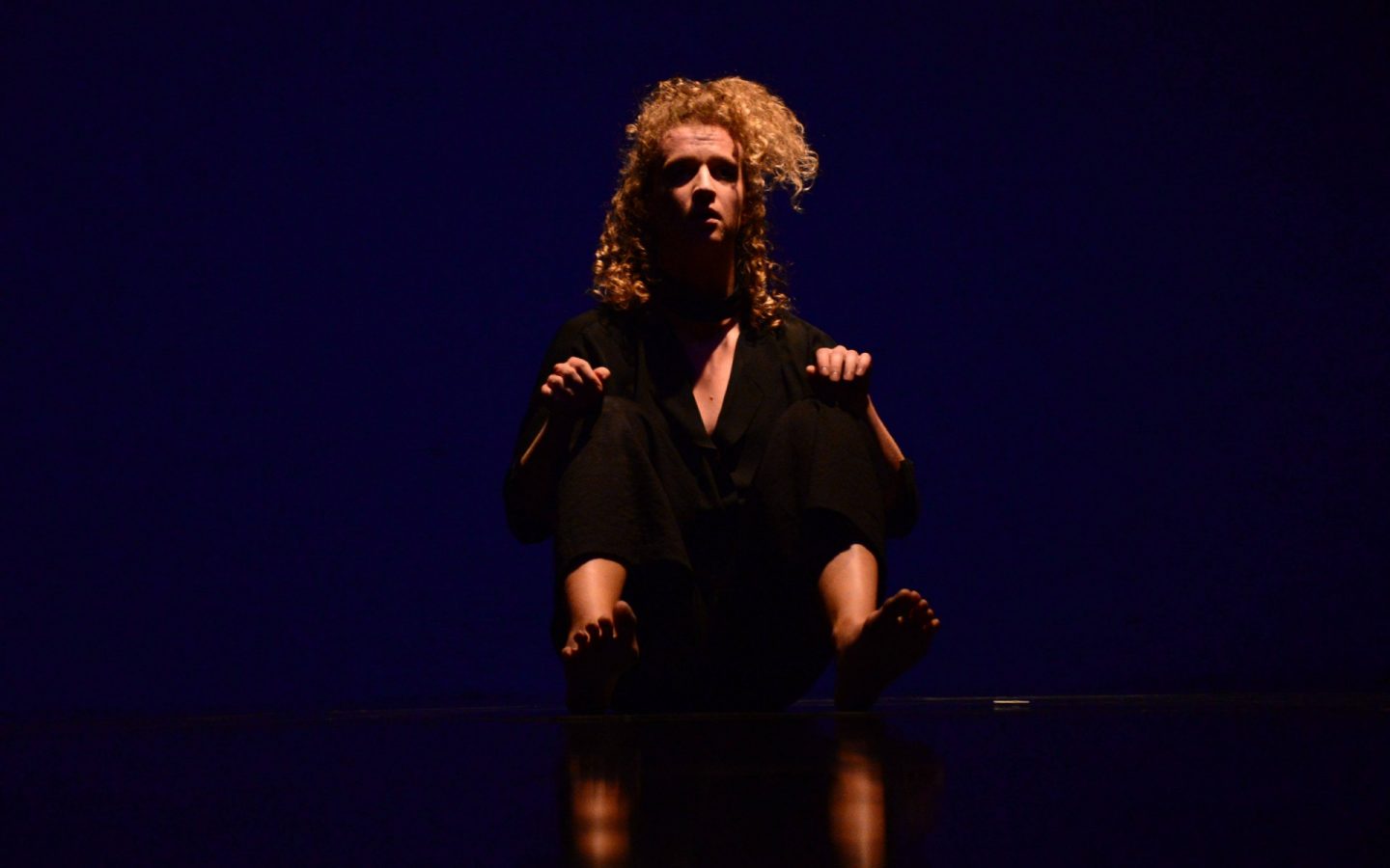 Zdjęcie. Na ciemnej scenie siedzi tancerka w czarnym kostiumie. Ma rozwiane włosy. Jest bosa.