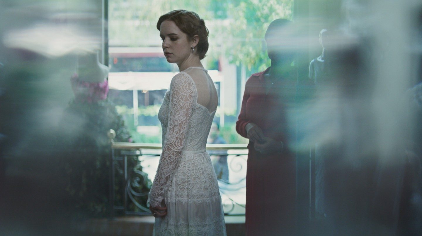 Kobieta w sukni ślubnej. Stoi we wnętrzu sklepu, w tle kolejne suknie na manekinach. Tło częściowo rozmyte.