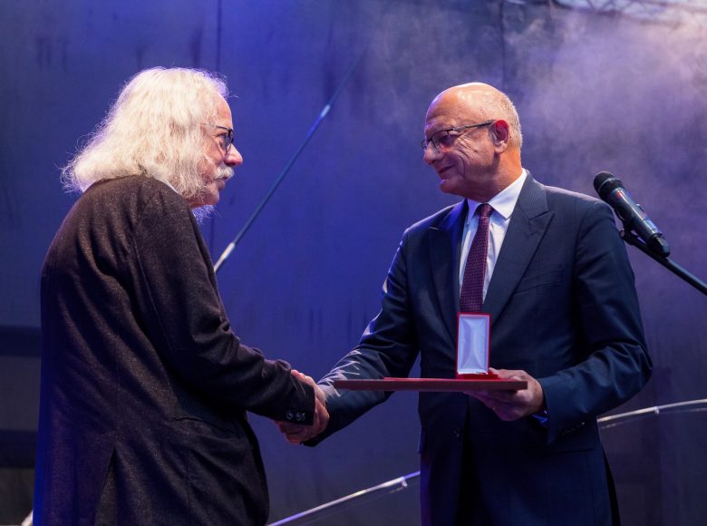 Zdjęcie przedstawia dwóch mężczyzn. Stoją na scenie. Jeden wręcza drugiemu medal i podaje mu dłoń w geście gratulacji.