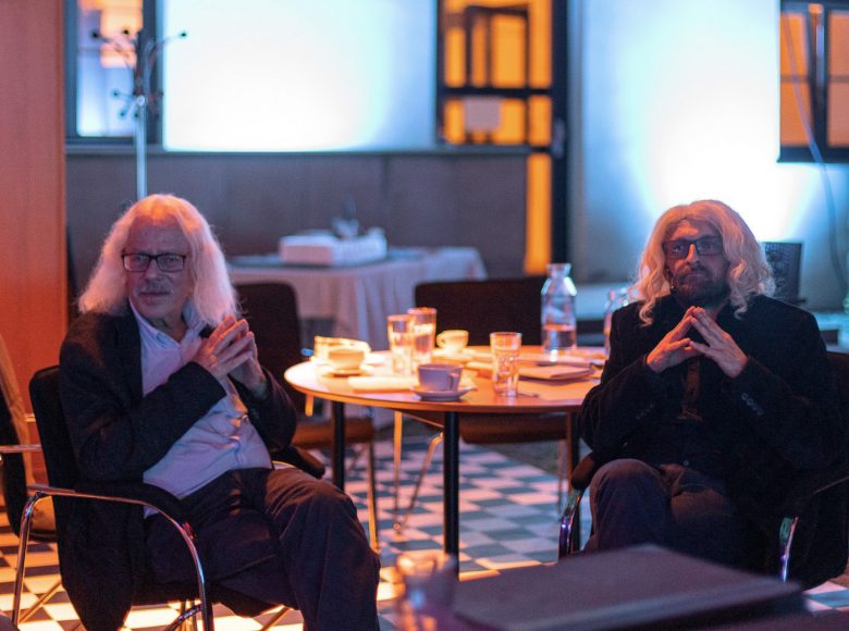 Zdjęcie. Zbliżenie na dwóch elegancko ubranych mężczyzn. Siedzą przy stoliku w takich samych pozach.