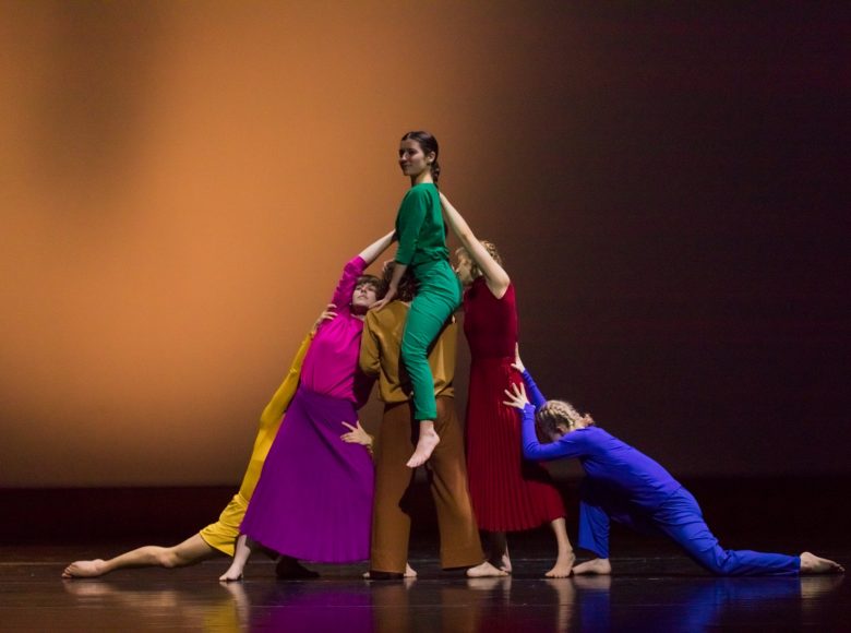 Zdjęcie. Grupa tancerek w kolorowych strojach znajduje się na scenie. Tworzą ze swoich ciał górę. Na jej szczycie siedzi tancerka w zielonym kostiumie.